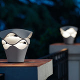 Cornet B/27 Cone LED bollard light in Stone Grey, tapered cone base with stacked wavy layers glowing white, shown outdoors on a wooden ledge at dusk.