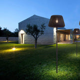 Ralph 50 outdoor floor lamp with woven cone shade in metallic brown, illuminated on a slim stem and tripod base on a lawn beside a stone house at dusk.