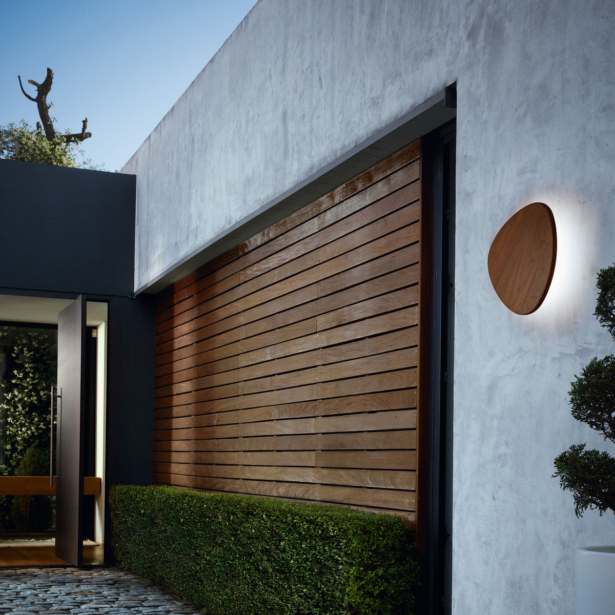 Tria 05 Wood LED outdoor wall light with a pebble-shaped teak wood face, mounted on a plastered house exterior and emitting a soft halo glow beside a wooden slatted wall.
