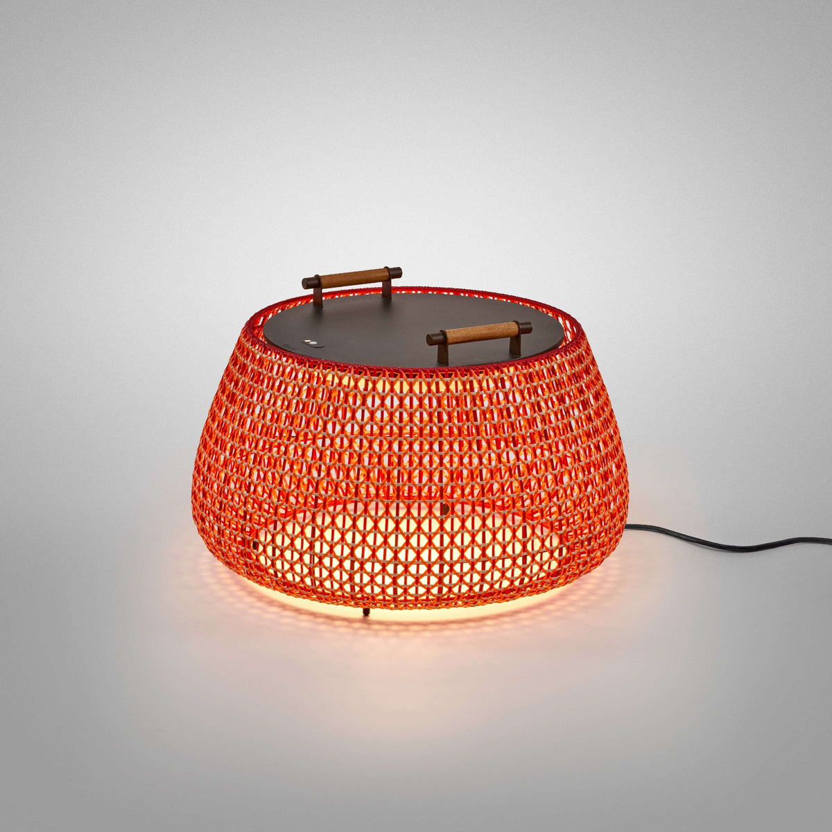 Nans Bag/38 LED outdoor floor lamp with a round woven red shade in an open lattice design, flat dark top with wood handles, glowing softly on a plain grey background.