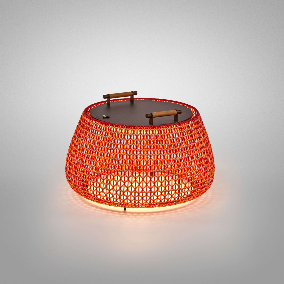 Nans Bag/38/R rechargeable outdoor floor lamp, wide woven red lattice basket shade with flat dark top and two teak handles, glowing LED light, on a plain grey background.