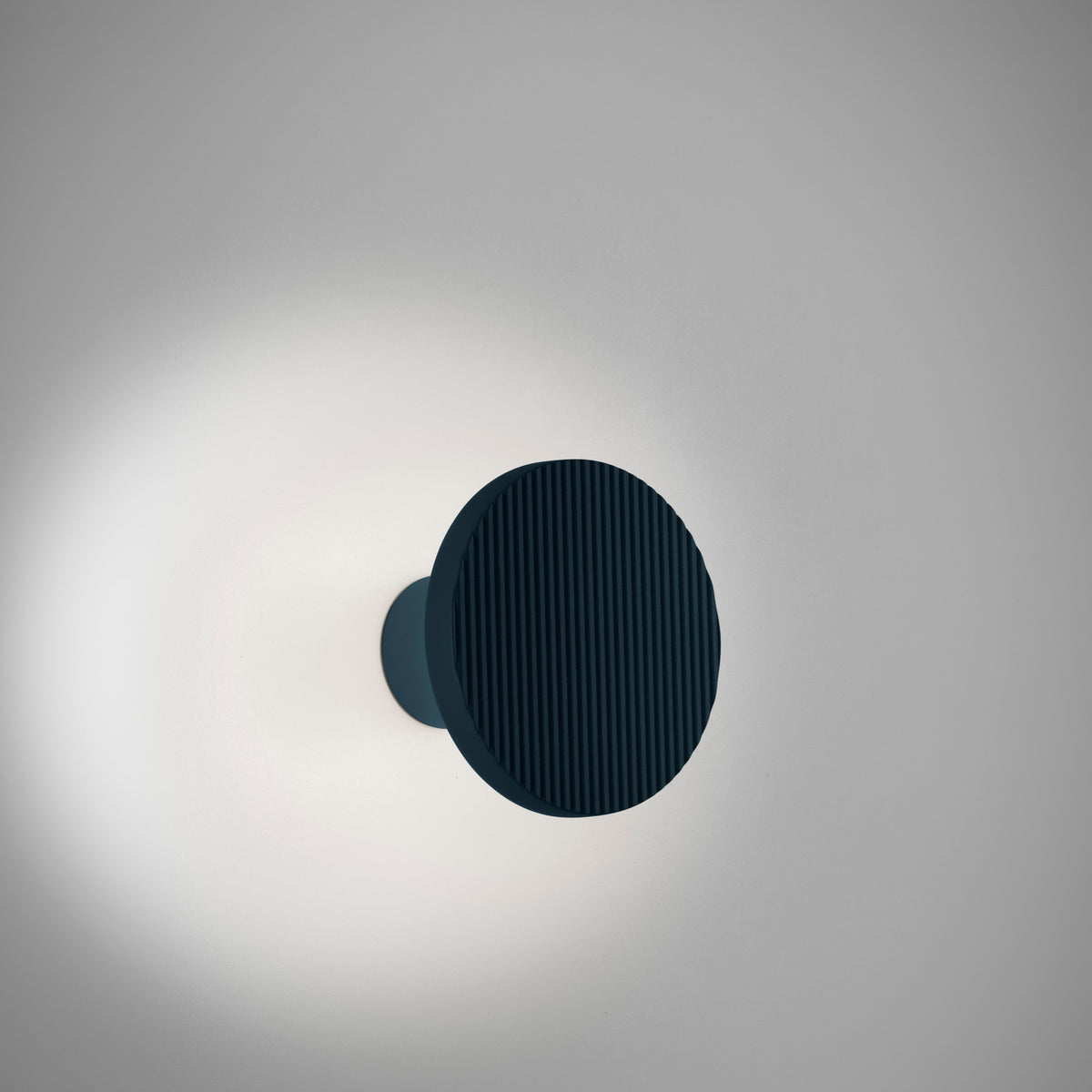 Arid A/01 round aluminium LED wall light with fluted disc in Textured Grey Blue, mounted on a short stem, casting a soft halo on a light grey background.