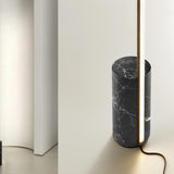 Hilow Line Nero Marquina floor lamp with a slim brushed brass LED rod rising from a black, white-veined marble cylindrical base, shown against a neutral interior wall.