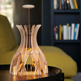 Domita M/36 wood bell table lamp with interlocking natural beech slats forming a flared shade, brown central stem and round knob, shown lit on a side table in a living room.