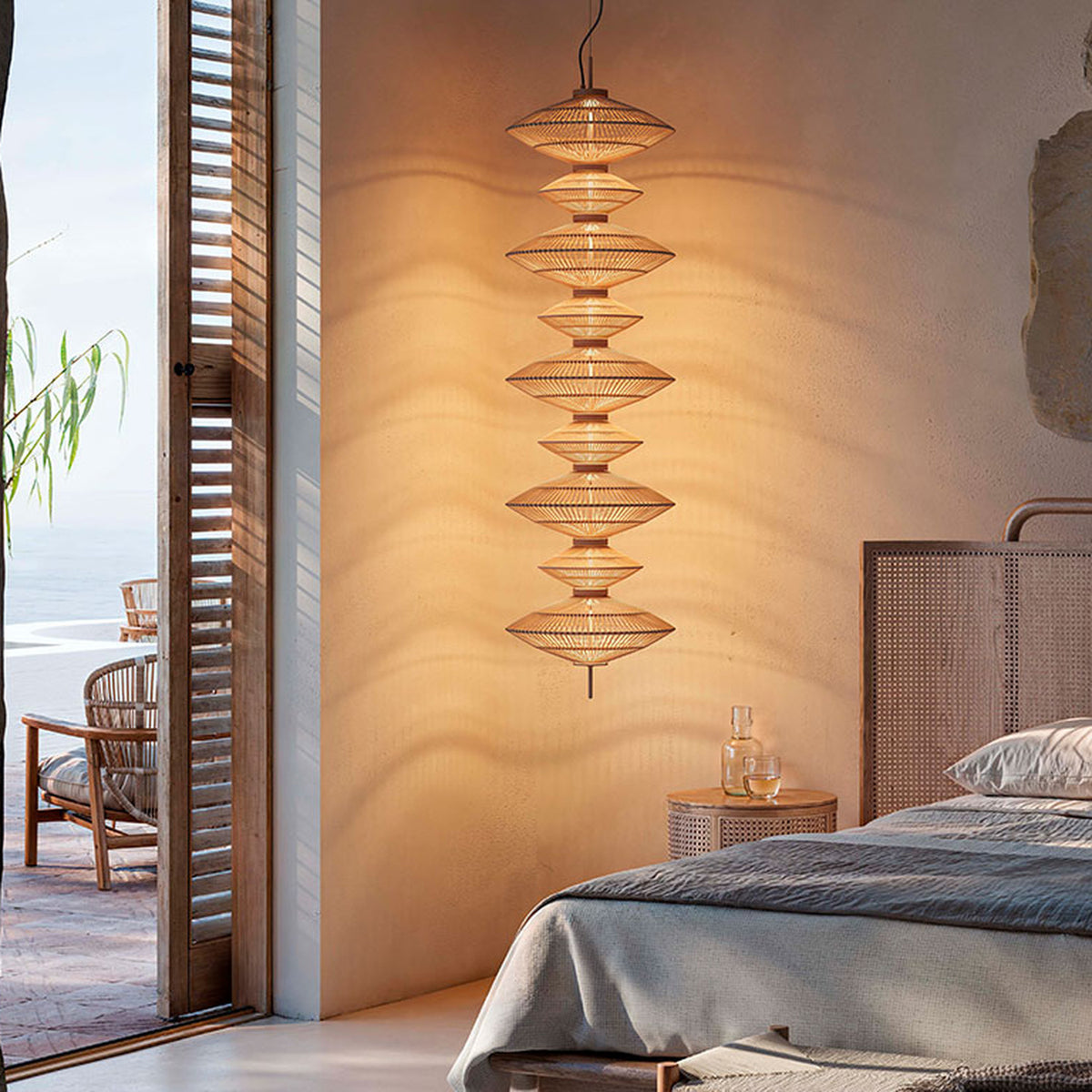 Yuyun S/9C LED pendant light with nine stacked woven cotton shades in satin beige, hung beside a bed in a bedroom with rattan headboard and bedside table.