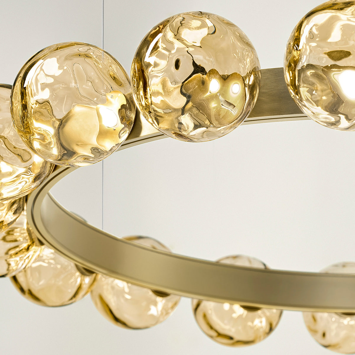 Close-up of the Murané R 25 ring chandelier showing a brushed champagne band set with textured caramel glass spheres, lit from within against a plain light background.
