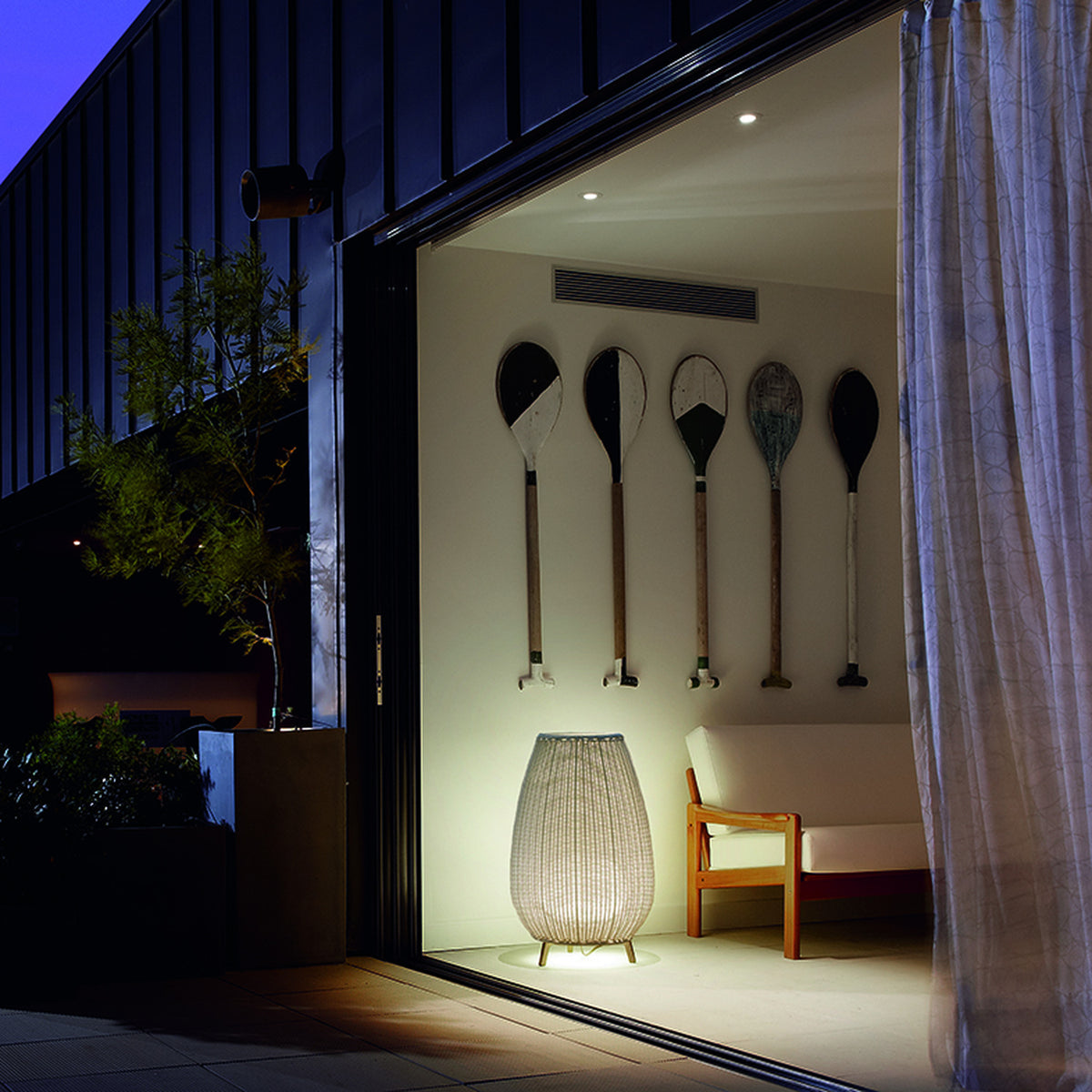 Amphora 01 Woven Linear LED Outdoor Floor Lamp in beige, lit on three short legs beside a sofa in a patio-style indoor-outdoor living space at night.