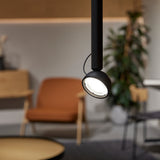 Berry S Aluminium LED Pendant Light with slim black tube and angled domed head, shown illuminated in a living room with an orange armchair and plant in the background.