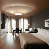 Siam 200 Ribbon Ceiling Light in cream, a wide pleated cone shade with white centre disc, shown illuminated above a bed in a bedroom setting.