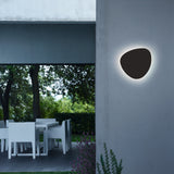Tria 04 Wood LED outdoor wall light with pebble-shaped graphite brown metal shade casting a warm ambient halo, mounted on an exterior wall near a dining terrace.