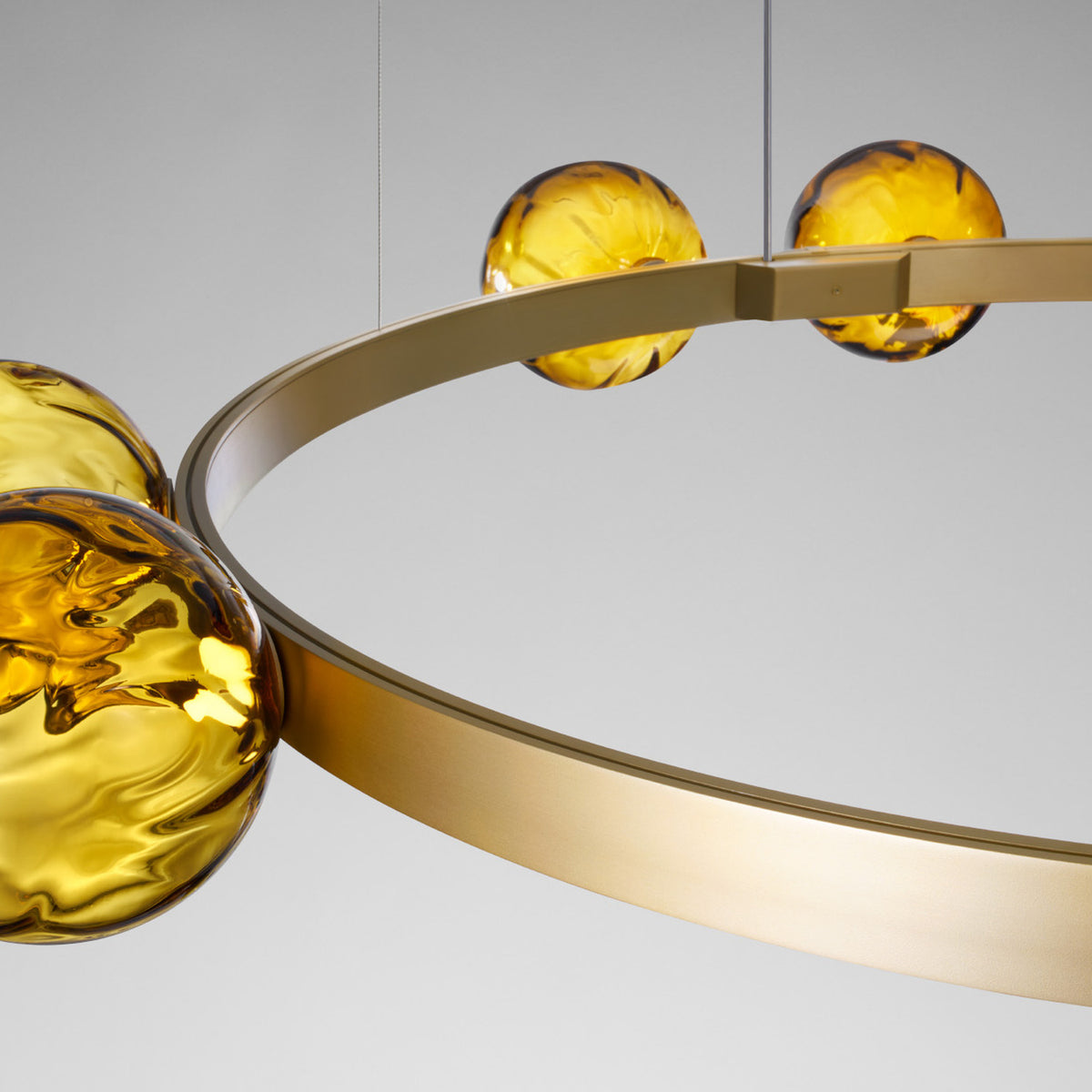 Murané R ring chandelier, close-up of brushed champagne band with paired amber glass spheres glowing warm gold, light grey background.