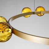 Murané R ring chandelier, close-up of brushed champagne band with paired amber glass spheres glowing warm gold, light grey background.
