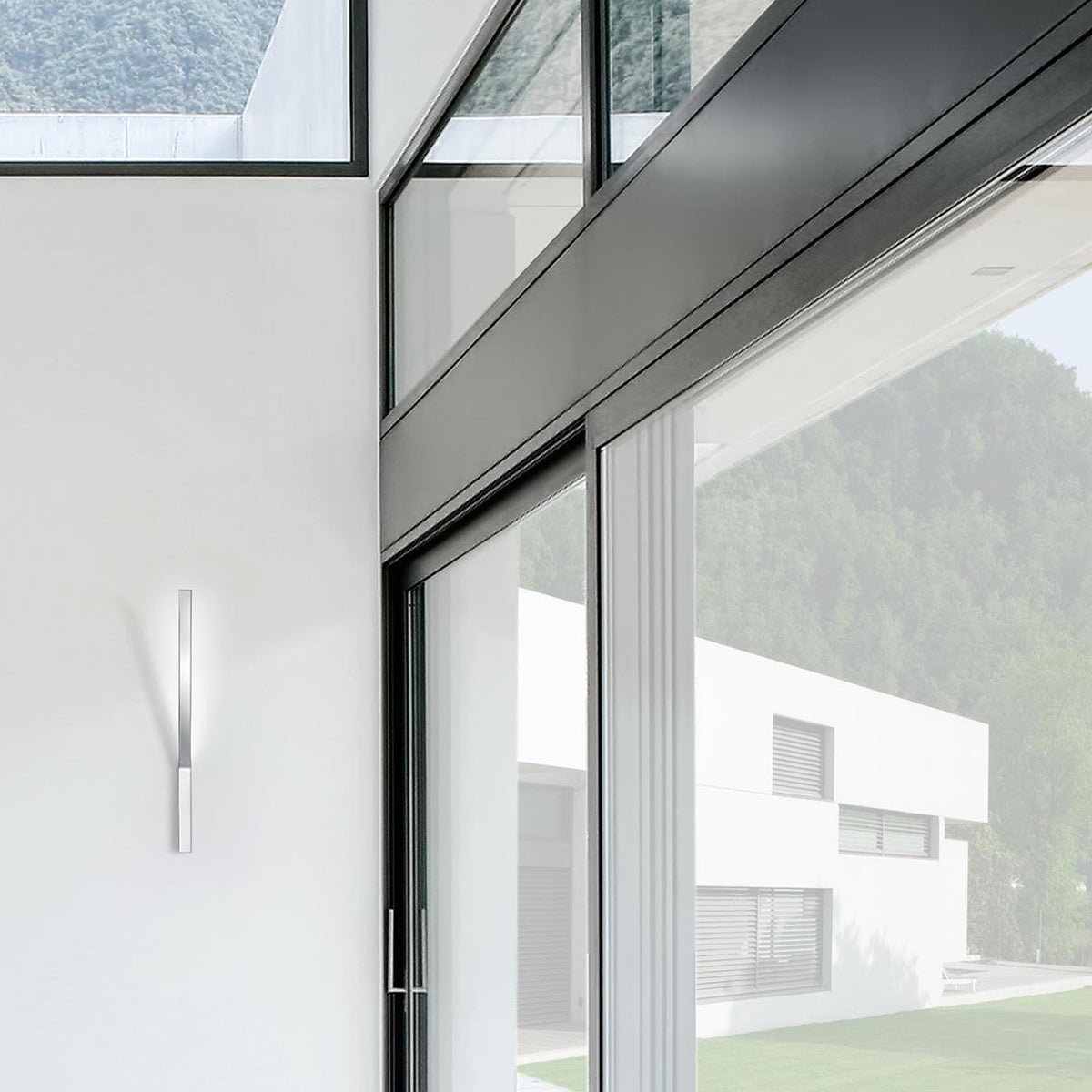 Ypsilon LED Wall Light in chrome, a slim vertical blade fixture mounted beside a sliding glass door in a modern white-walled interior with hillside views.