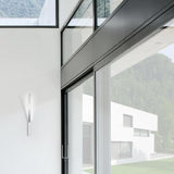 Ypsilon LED Wall Light in chrome, a slim vertical blade fixture mounted beside a sliding glass door in a modern white-walled interior with hillside views.