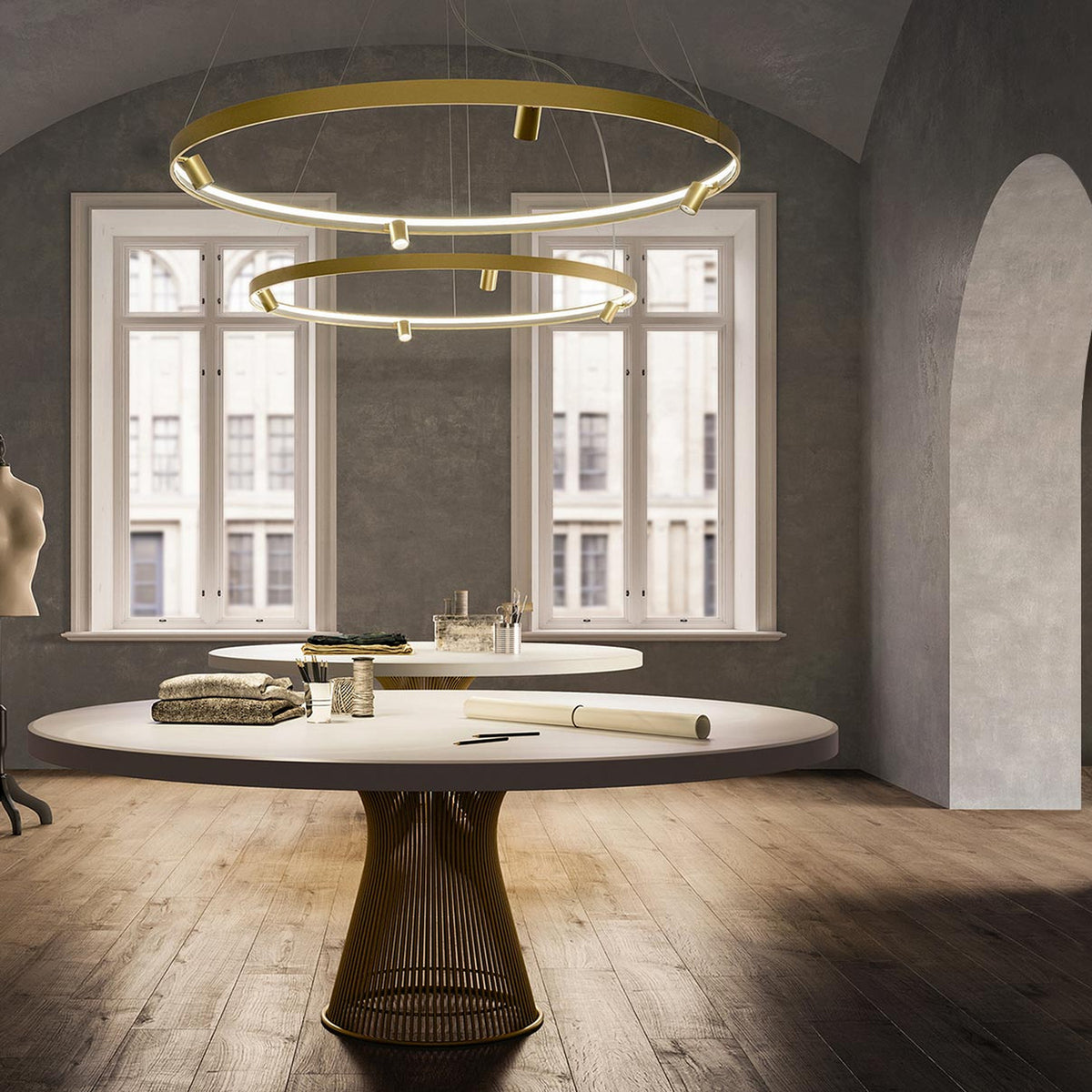Arena Aluminium Ring LED Pendant Light in brass finish, two tiered circular hoops with integrated LED and small spotlight heads, shown above a dining table in a neutral-toned room.