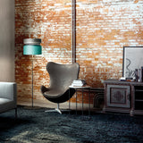 Aspen F40 Brass Drum Floor Lamp with grey and teal stacked drum shade on a slim stem base, shown beside an armchair in a living room with exposed brick wall.