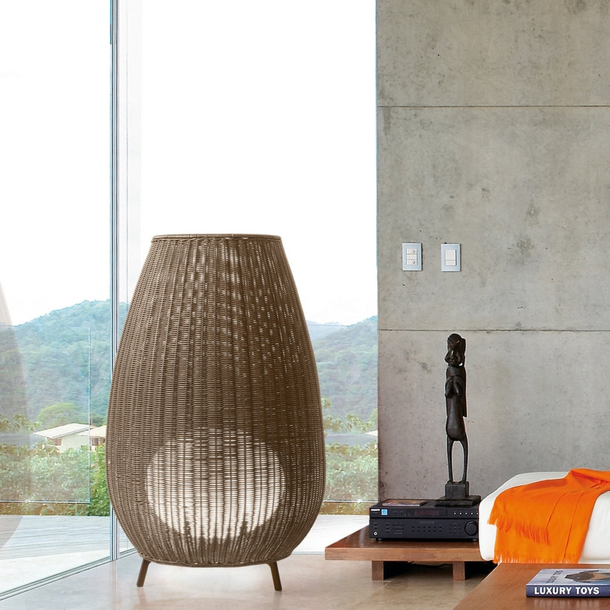 Amphora 03 Rattan LED Outdoor Floor Lamp in brown, tall woven amphora-shaped shade on three legs, shown beside floor-to-ceiling windows in a modern living room.