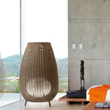 Amphora 03 Rattan LED Outdoor Floor Lamp in brown, tall woven amphora-shaped shade on three legs, shown beside floor-to-ceiling windows in a modern living room.
