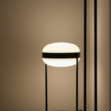 Beaded T Glass LED table lamp with a rounded opal white shade held by a dark centre band between slim black rods, glowing warmly in a softly lit room.