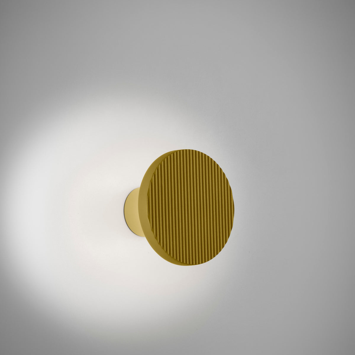 Arid A/01 round aluminium LED wall light with ribbed sand yellow disc on a short stem, casting a soft ambient glow against a plain light grey background.