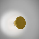 Arid A/01 round aluminium LED wall light with ribbed sand yellow disc on a short stem, casting a soft ambient glow against a plain light grey background.