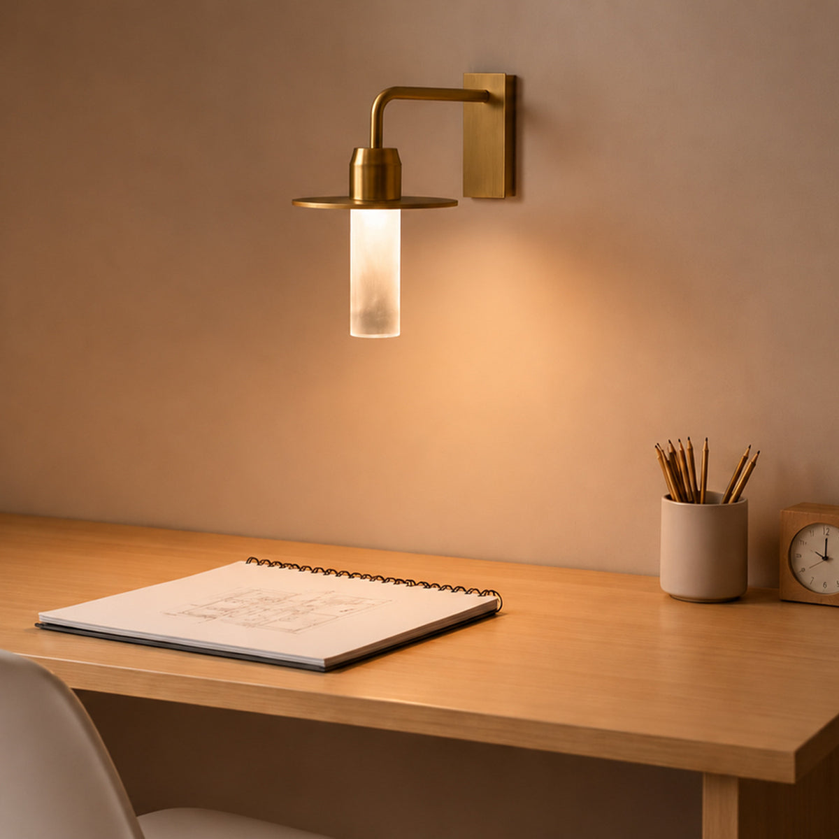 Beira W Glass LED Wall Light in brushed brass, with a rectangular wall plate, downturned arm, round disc and frosted diffuser tube, shown lit above a wooden desk.