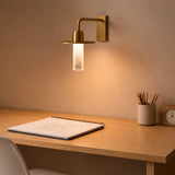 Beira W Glass LED Wall Light in brushed brass, with a rectangular wall plate, downturned arm, round disc and frosted diffuser tube, shown lit above a wooden desk.