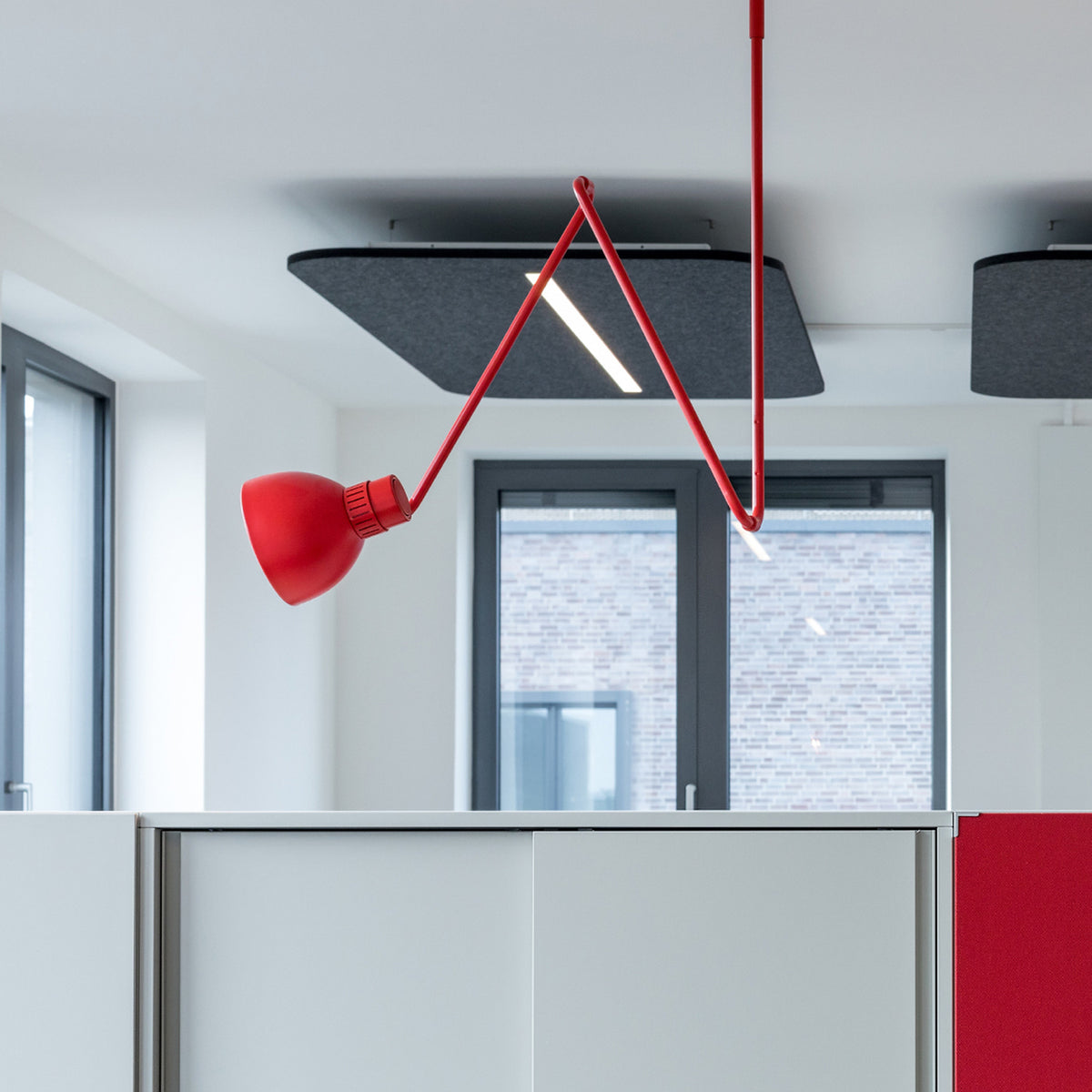 Blux C Aluminium Dome Ceiling Light in red, with angled zig-zag arms and a round dome shade offset to one side, installed above an office partition with large windows behind.