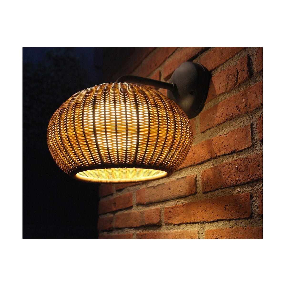 Garota A/01 Globe LED Outdoor Wall Light in Brown, a woven flattened globe shade on a curved arm with round wall mount, mounted on a brick wall at night.