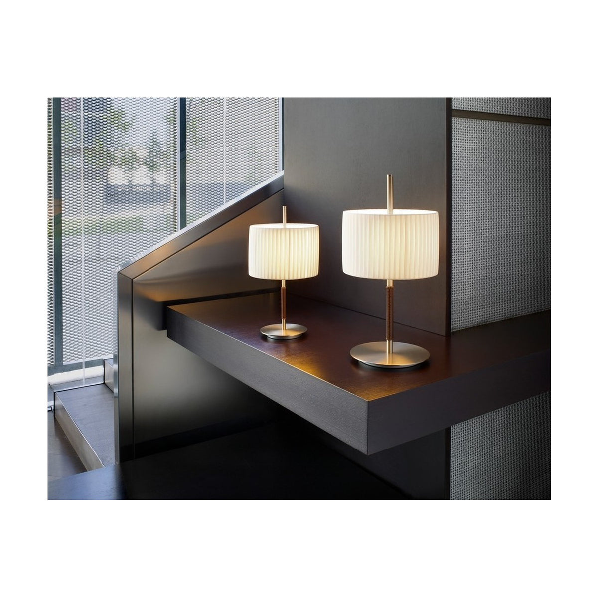 Two Danona Mini table lamps with pleated white ribbon drum shades on slim satin nickel stems and round bases, shown on a dark wood shelf in a modern interior.