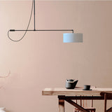 Raisa Fabric Drum Pendant Light in matt black, with a slim offset arm and white fabric drum shade, shown above a wooden dining table in a pink-toned room.