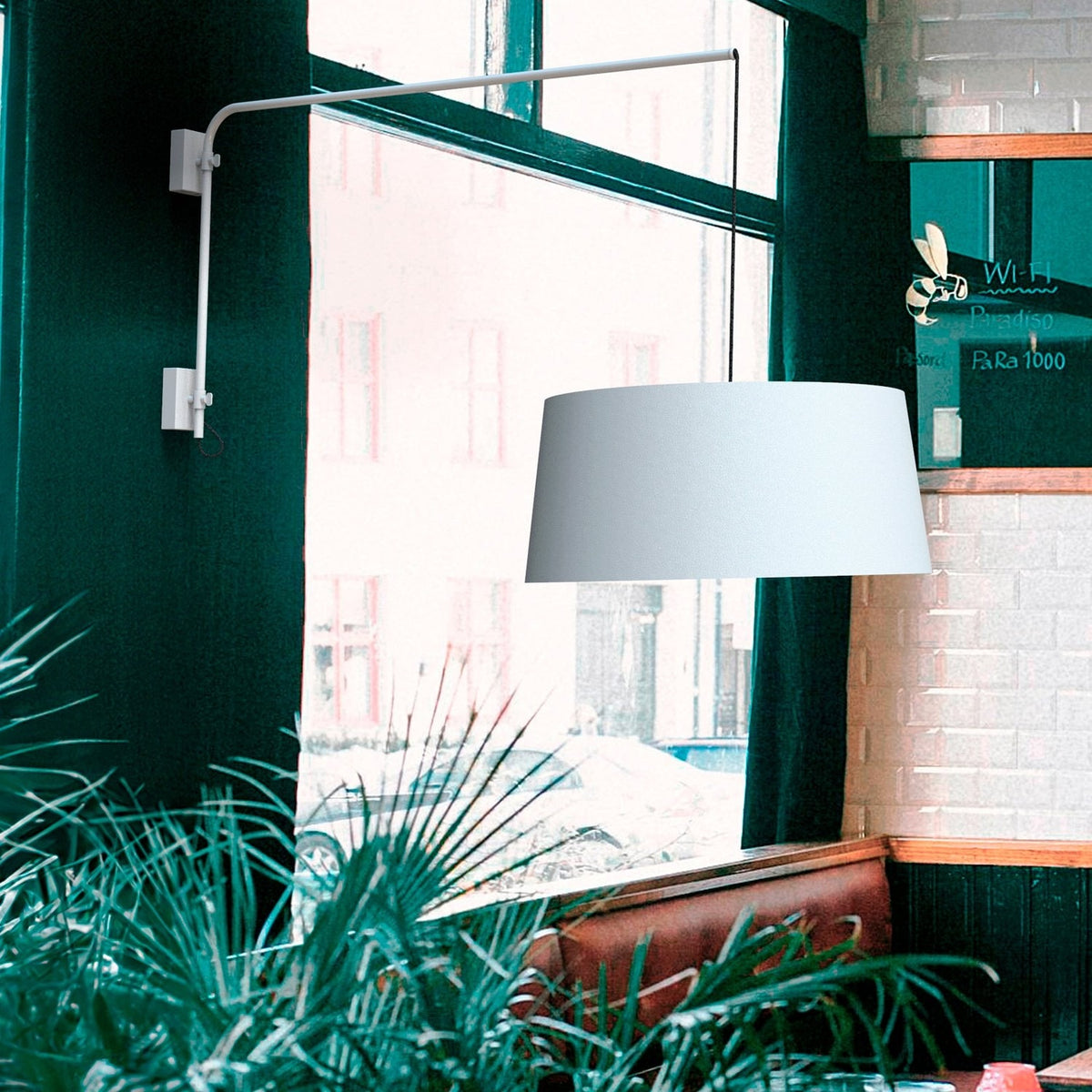 Raisa swing arm wall light in matt white, long adjustable arm extending from a wall bracket with a white fabric drum shade suspended on a cable, shown near a window in a plant-filled dining area.
