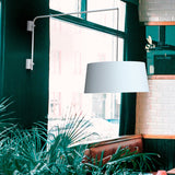 Raisa swing arm wall light in matt white, long adjustable arm extending from a wall bracket with a white fabric drum shade suspended on a cable, shown near a window in a plant-filled dining area.