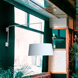 Raisa Fabric Drum Adjustable Swing Arm Wall Light in matt white, with a long swing arm and fabric drum shade on a textile cable, shown in a dark green café interior near a window.