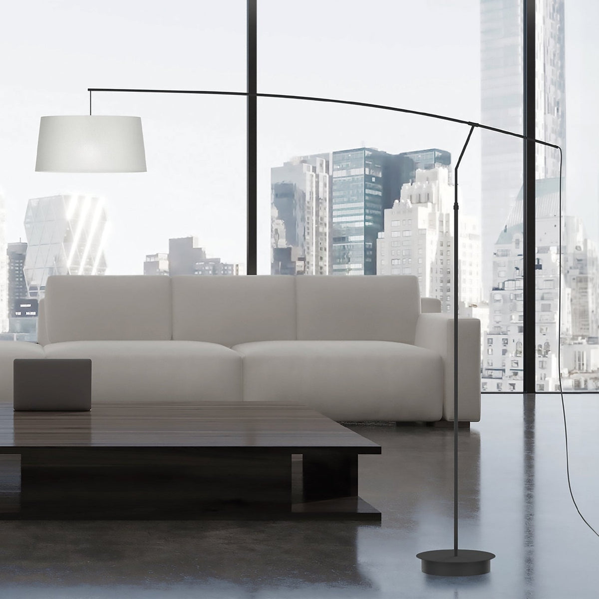 Raisa Fabric Drum Arc Floor Lamp in matt black, with a slim iron stem and curved overarching arm holding a white fabric drum shade, shown beside a sofa in a city-view living room.