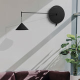 Roma Cone Wall Light in matt black, with round disc backplate, slim extending arm, wire support, and conical shade, shown mounted in a living room setting.