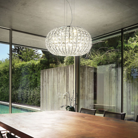 In a contemporary dining room featuring a spacious wooden table and chairs, the Auroral 5 Light Crystal Suspension in Chrome elegantly illuminates the space. Floor-to-ceiling glass windows offer views of a lush garden and pool beyond, with an orchid as the table's centerpiece.