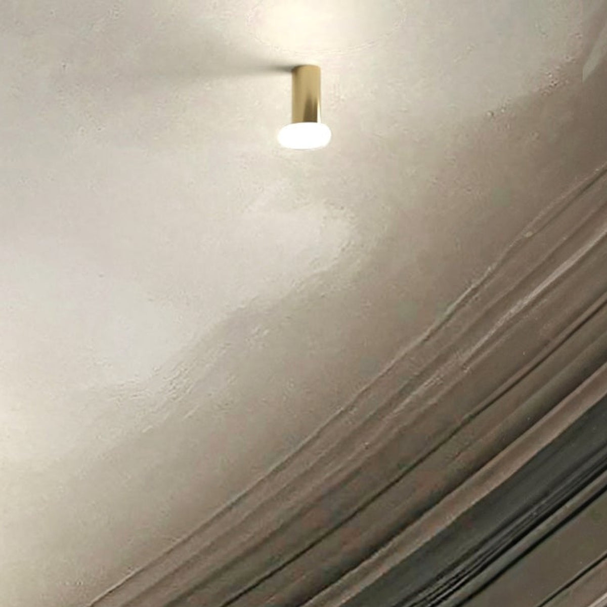 Rina ceiling-mounted downlight with slim polished brass cylinder and round opal glass diffuser, glowing softly against a textured light grey ceiling.