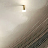 Rina ceiling-mounted downlight with slim polished brass cylinder and round opal glass diffuser, glowing softly against a textured light grey ceiling.