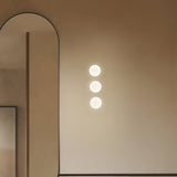 Three Rina opal glass recessed wall lights in polished brass mounted vertically on a tan wall beside an arched mirror, each glowing softly.