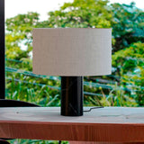 Romy Drum Table Lamp with a wide beige cotton drum shade on a black marble cylindrical base, shown outdoors on a wooden surface with greenery behind.