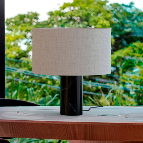 Romy Drum Table Lamp with a wide beige cotton drum shade on a black marble cylindrical base, shown outdoors on a wooden surface with greenery behind.