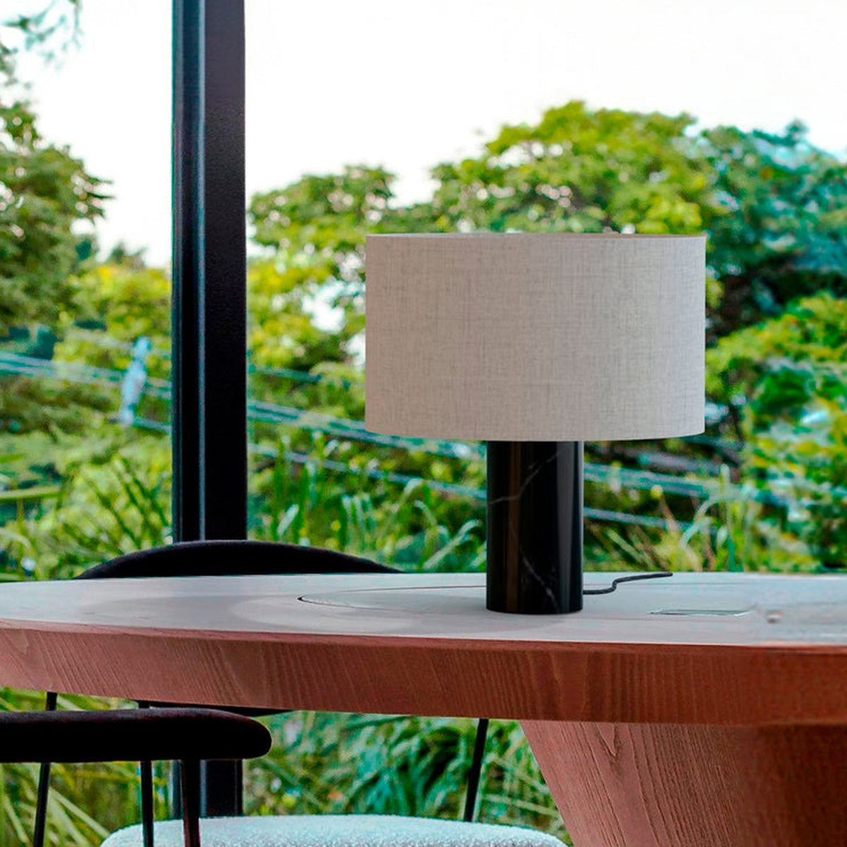 Romy Drum Table Lamp with a black marble cylindrical base and beige cotton drum shade, shown on a wooden table against a greenery-filled window backdrop.