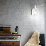 A cozy room corner with a wooden side table holding a vintage radio, a dark bottle, and a bowl of green apples. The minimalist design includes an auburn wall light in white casting soft, ambient lighting on the textured gray wall. Nearby, a black chair with a fluffy cream throw invites relaxation.