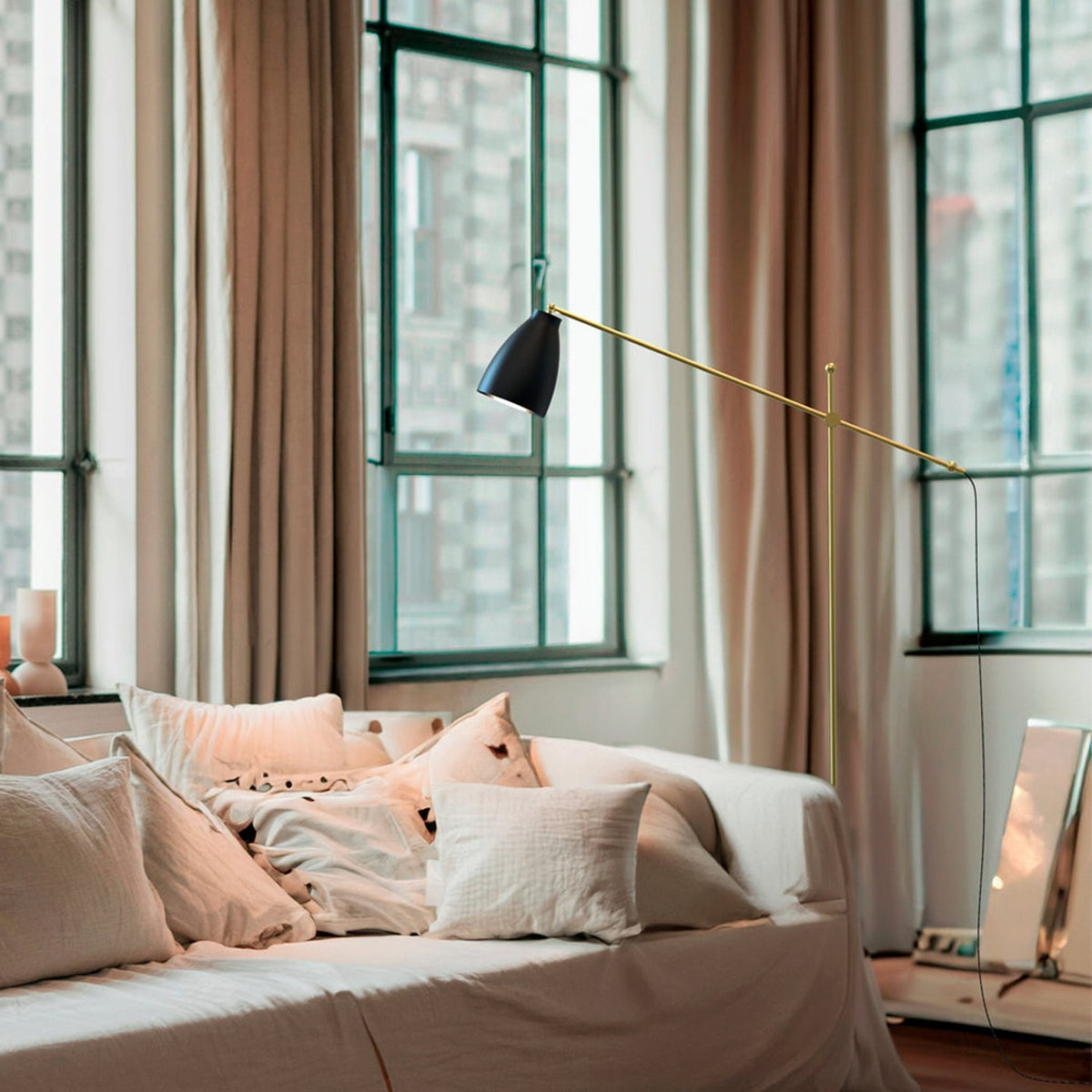 Nala Adjustable Floor Lamp with Marble Base in matt black and gold, angled over a bed with cushions in a bedroom with tall curtained windows.