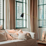 Nala Adjustable Floor Lamp with Marble Base in matt black and gold, angled over a bed with cushions in a bedroom with tall curtained windows.