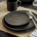 Stacked Neri stoneware side plates in black with ridged, fluted surfaces, set on a woven placemat with a dark mug and cutlery alongside.