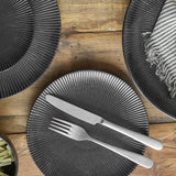 Neri Stoneware Side Plate in black with ribbed sunburst texture, set with a knife and fork, shown alongside matching plates on a wooden table.