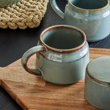 Pixie Stoneware Cup 350ml in green with a glossy reactive glaze and open loop handle, shown on a wooden board beside a matching jug and lidded pot.