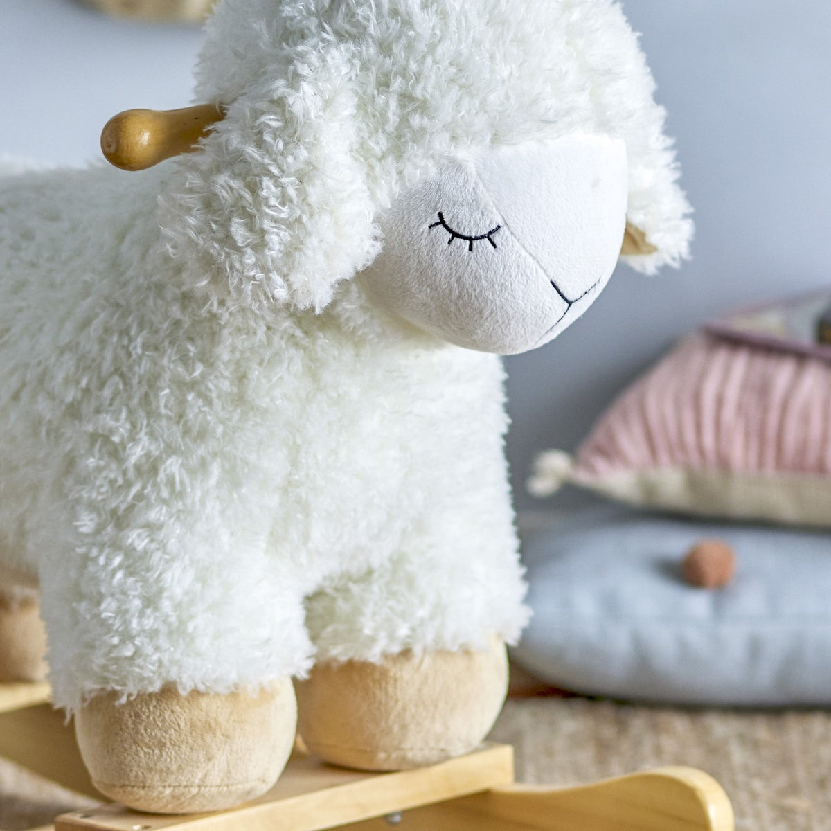 Close-up of the Laasrith Rocking Sheep in white, a curly fabric ride-on toy with a sleepy embroidered face, floppy ears, wooden handle, and suede-style hooves on wooden rockers.